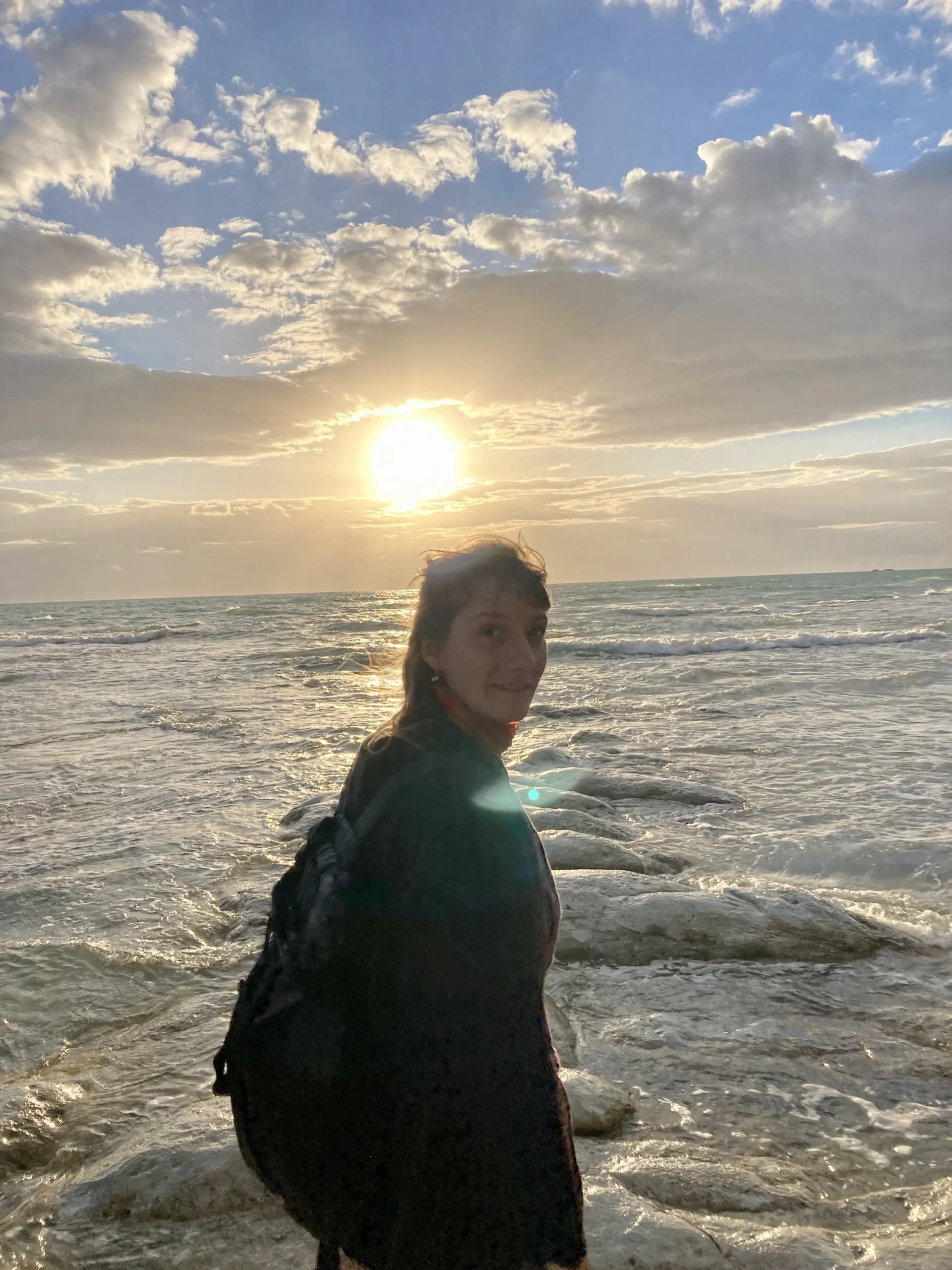 Ingrid Szilagyi steht auf einen Felsen umgeben von Wasser. Das offene Meer, im Hintergrund ist die Sonne am untergehen. Sie schaut in die Kamera.